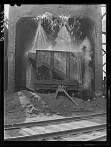 A black and white photo of an industrial-sized container of hot ashes being soaked by water from spigots above