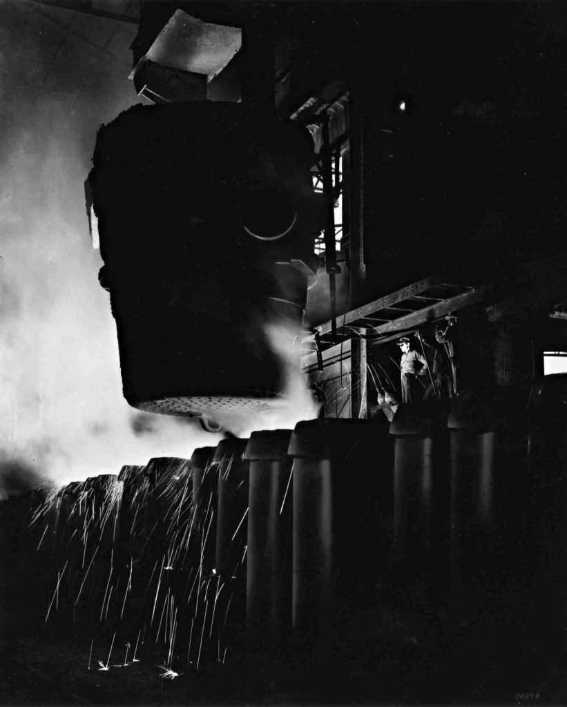 A black and white photo shows a worker standing next to an en enormous ladle of molten metal