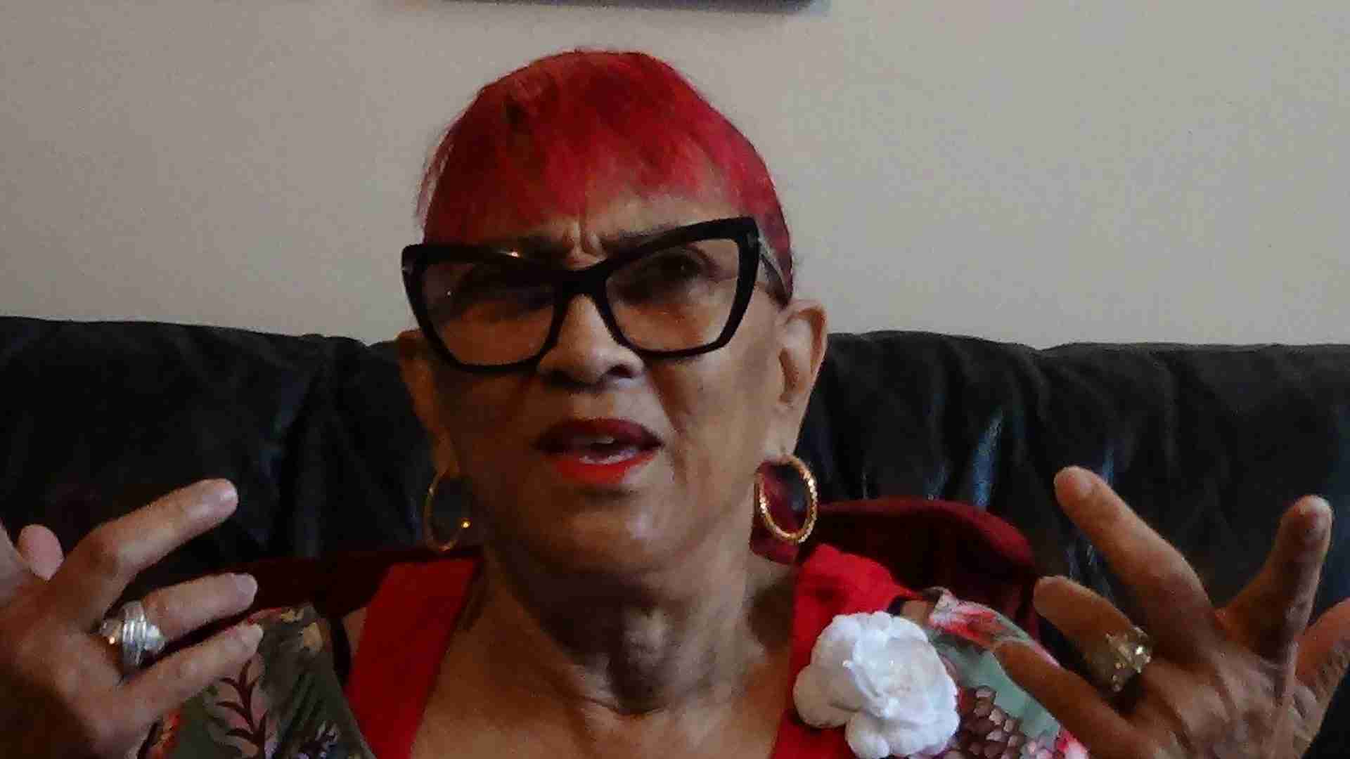 A woman with dyed red hair speaks and gestures with her hands.