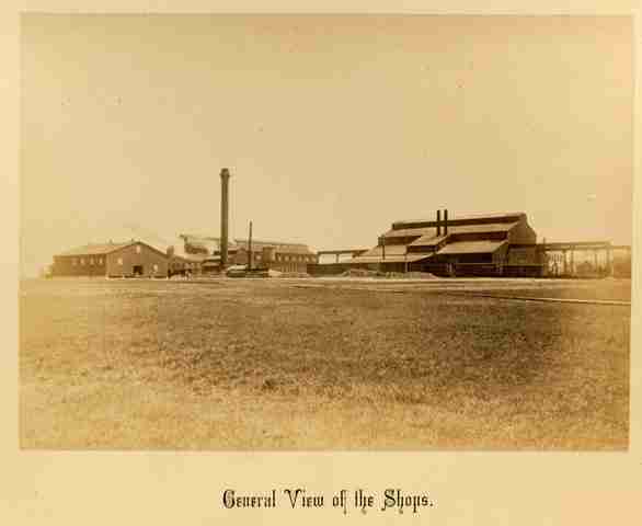 A sepia tone photograph shows a few industrial buildings surrounded by fields