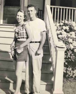 A black-and-white photo shows a young couple outside a suburban house in the 1950s