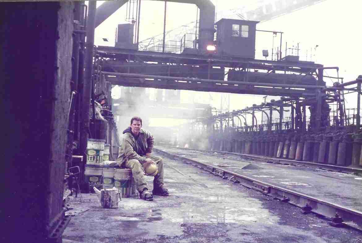 A color photograph shows a man seated on the roof of a large industrial building wearing work clothes.