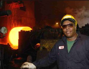 A color photograph shows molten, liquid steel being shaped in heavy machinery with a Black worker wearing a jumpsuit, helmet, gloves, and goggles.