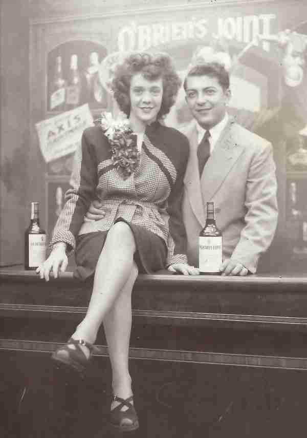 A black and white photograph shows a young woman sitting on a bar counter next to a young man in a suit.