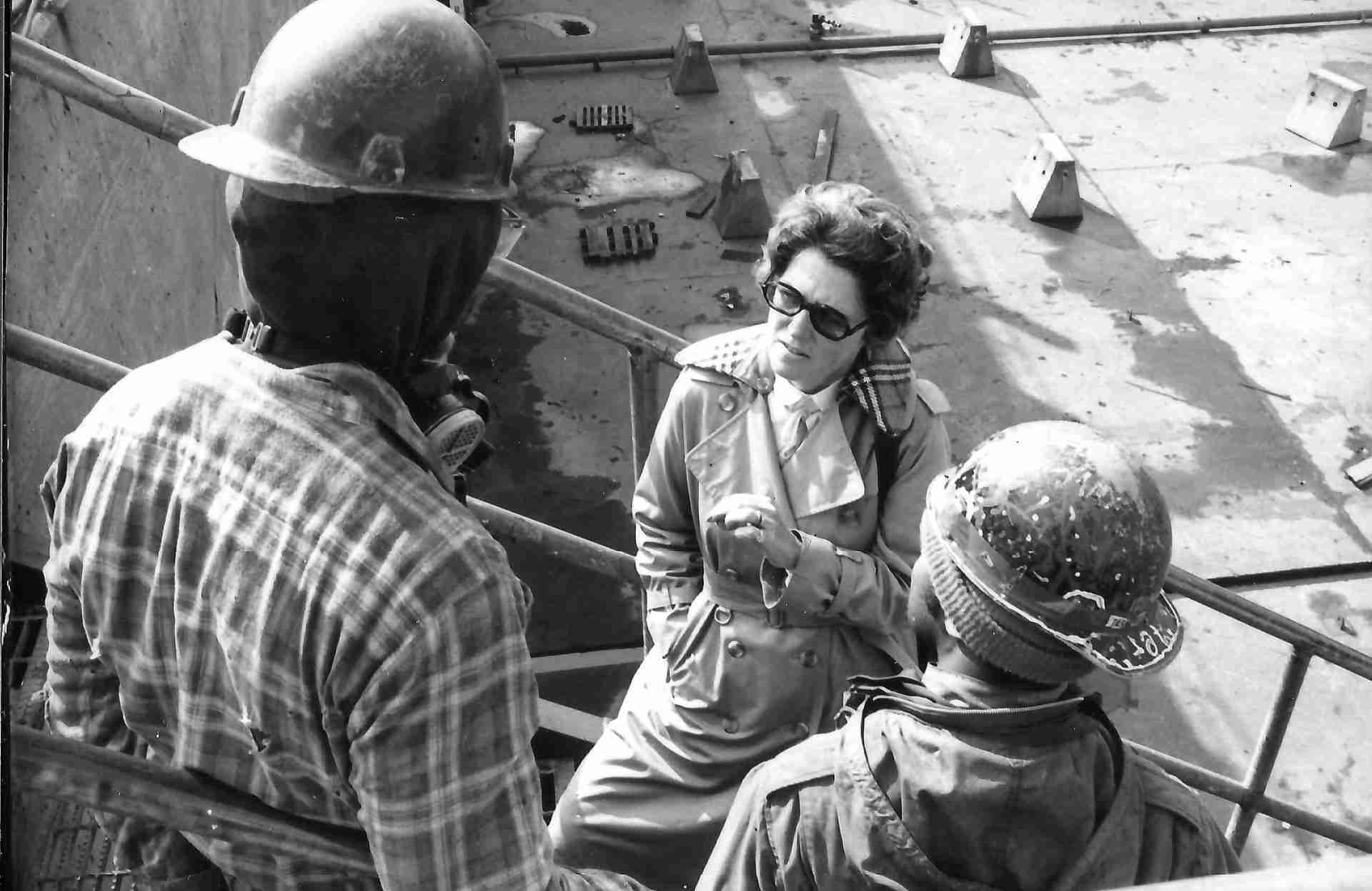 A black and white photo from 1982 shows a woman meeting with two shipyard workers on an exterior staircase
