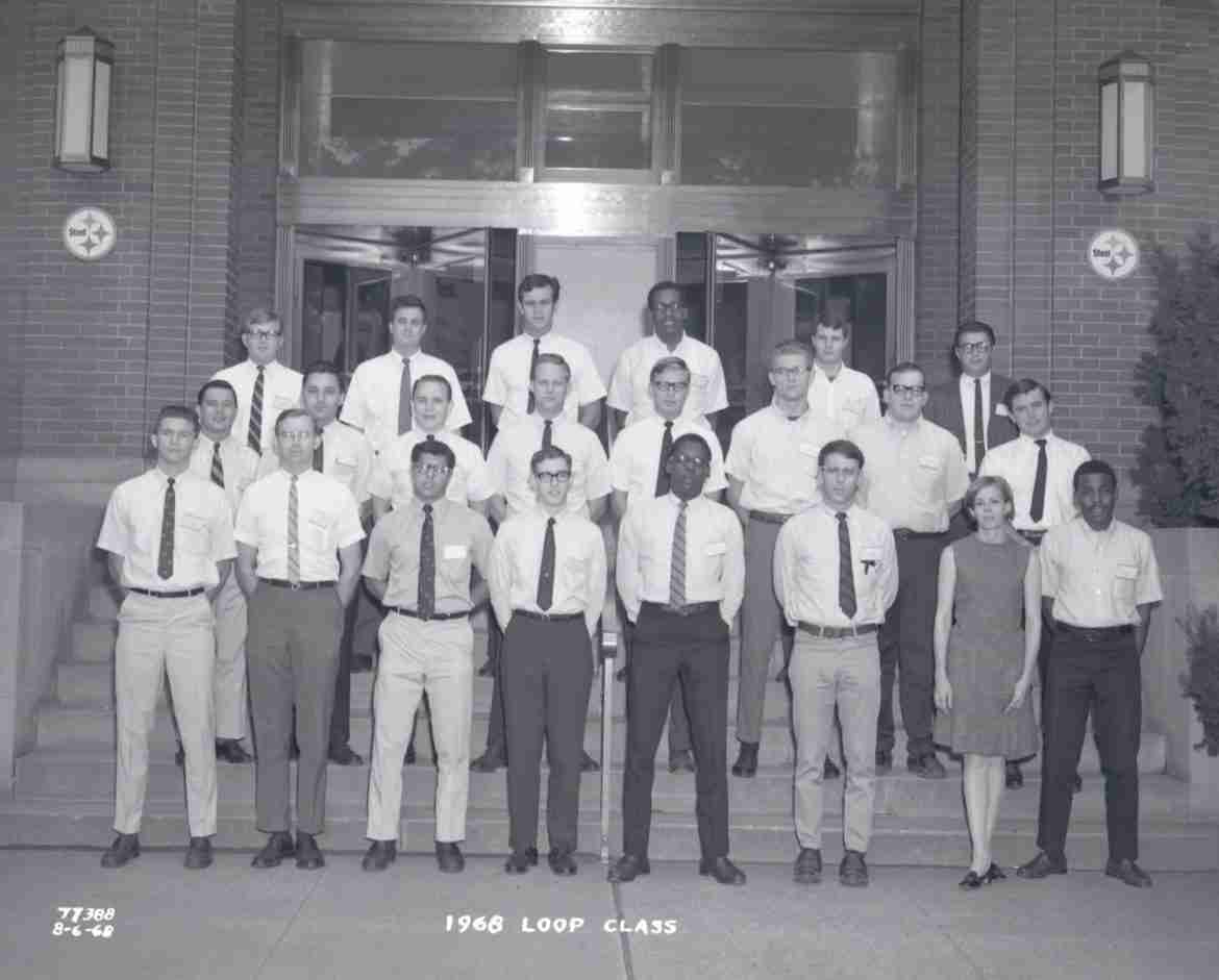 A black and white photo shows a group of young engineers wearing slacks and button-up shirts, standing on steps