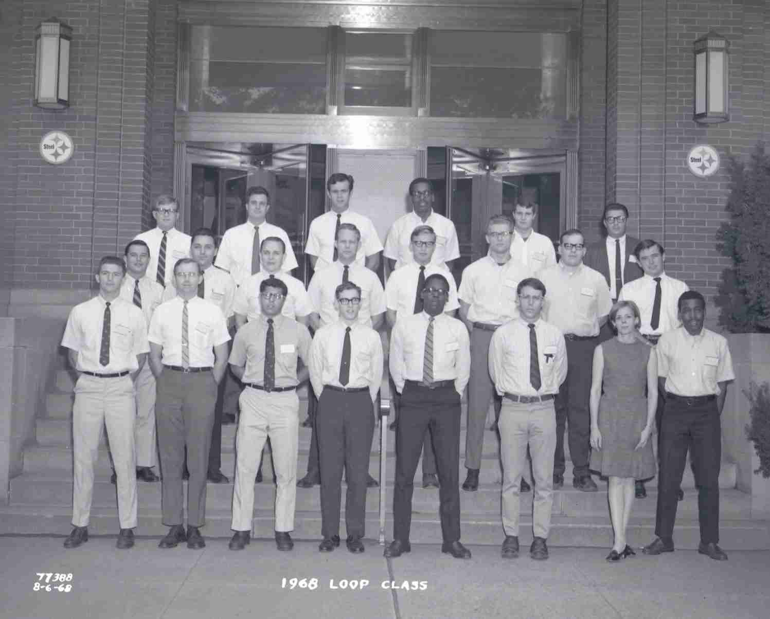 A black and white photo shows a group of young engineers wearing slacks and button-up shirts, standing on steps