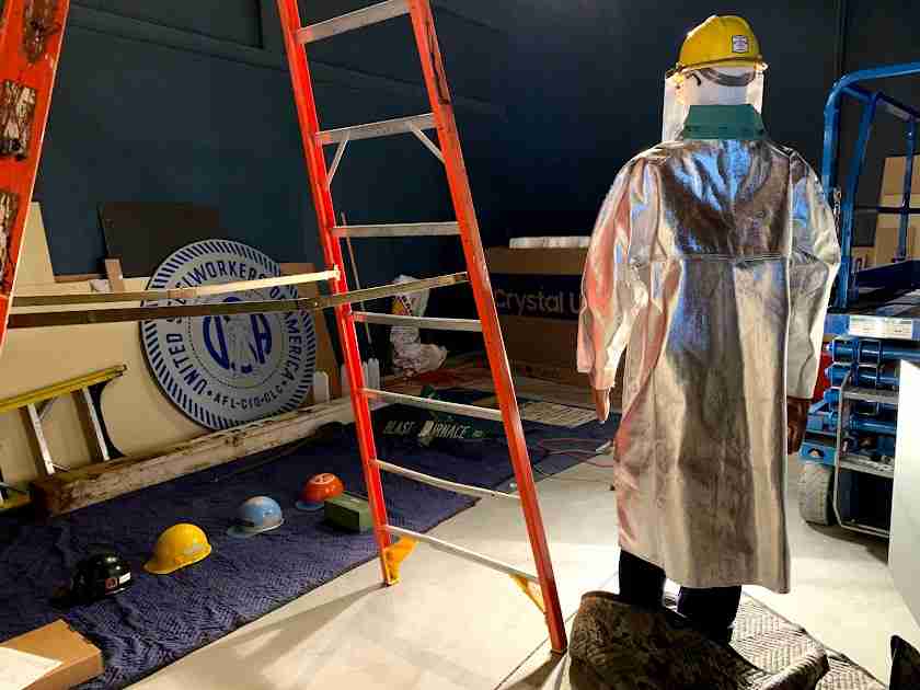 Parts of an exhibition taking shape: a mannequin wearing a fire-resistant silver coat and hard had, a ladder, work helmets, and other signage