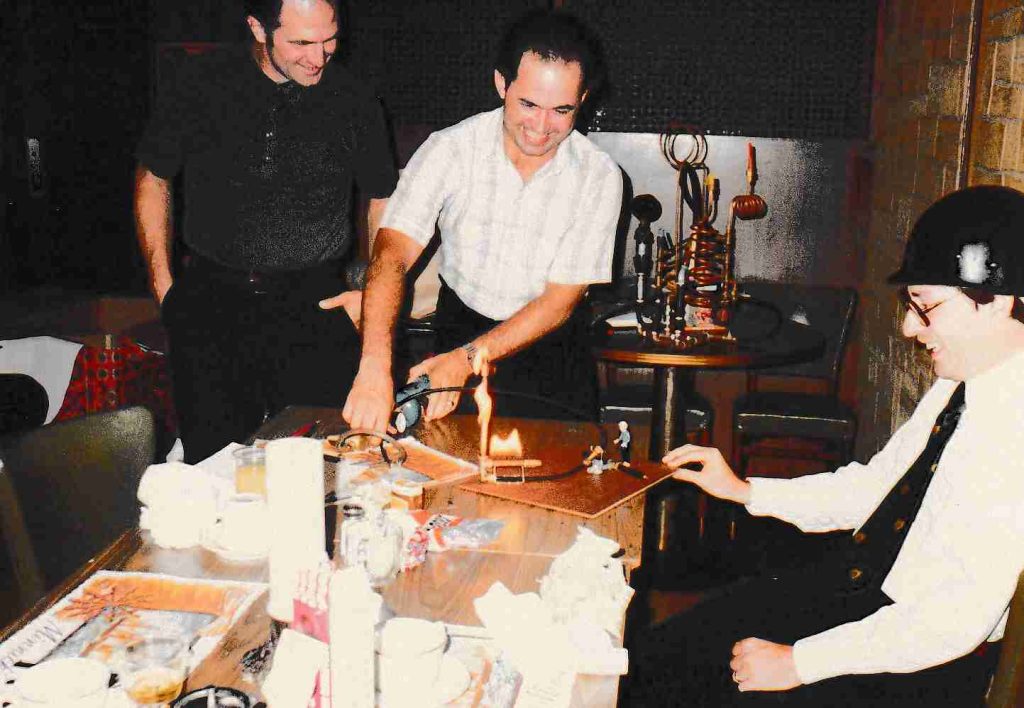 A color photo shows three men smiling and looking at a small, working model of the L blast furnace on a table with food and drinks