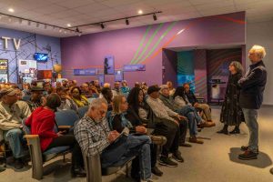 A small theater is crowded with people facing two producers, standing 